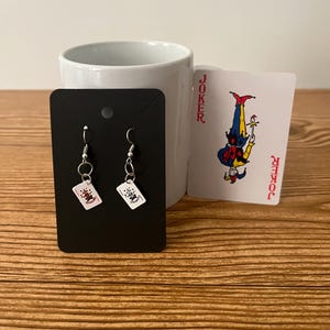 May include: A pair of silver earrings with small playing card charms. The charms are red and white and feature a joker design. The earrings are displayed on a black card with a white background.
