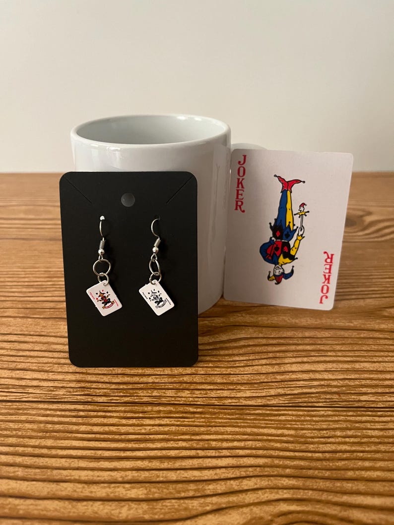 May include: A pair of silver earrings with small playing card charms. The charms are red and white and feature a joker design. The earrings are displayed on a black card with a white background.
