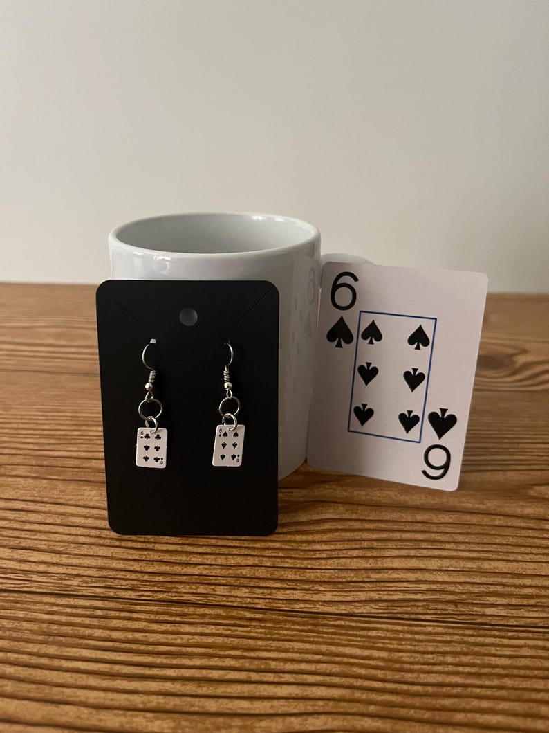 May include: A pair of silver earrings with a small playing card charm. The card is a six of spades.