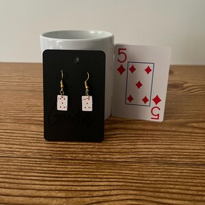 May include: A pair of gold earrings with a white card design featuring red diamonds and the number 5. The earrings are displayed on a black card with the word "Goodies" printed on it.