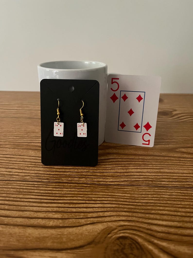 May include: A pair of gold earrings with a white card design featuring red diamonds and the number 5. The earrings are displayed on a black card with the word "Goodies" printed on it.