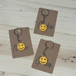 May include: Three yellow smiley face keychains with silver key rings are displayed on brown cardboard tags. The tags have the word "Goodies" written on them in a cursive font. The background is a light wood surface.