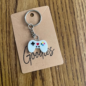 May include: A white video game controller keychain with a silver keyring. The controller has a black cross, red heart, and colorful buttons. The keychain is attached to a brown card with the word "Gooques" in cursive.