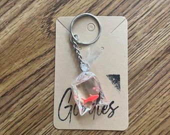 May include: A keyring featuring a miniature clear plastic bag filled with liquid and a small orange fish charm. The keyring is attached to a silver metal ring and chain, mounted on a rectangular tan card with the word Giggles printed in black.
