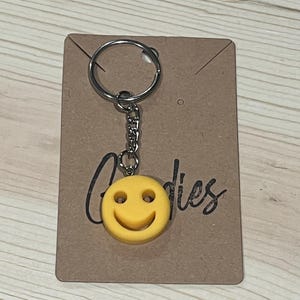 May include: A yellow smiley face keychain with a silver chain and keyring. The keychain is attached to a brown cardboard tag with the word "Goodies" in a handwritten font.