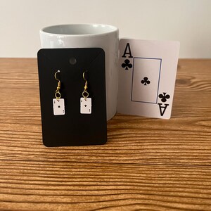 May include: A pair of gold dangle earrings with a white card design featuring the Ace of Clubs. The earrings are displayed on a black card with a white border.