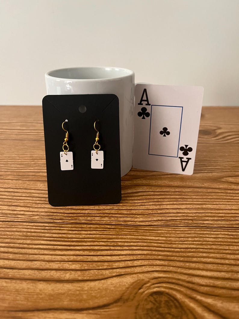 May include: A pair of gold dangle earrings with a white card design featuring the Ace of Clubs. The earrings are displayed on a black card with a white border.