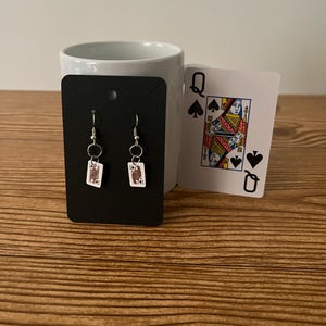 May include: A pair of silver earrings with a small white card featuring the Jack of Spades playing card design.