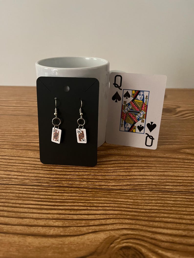 May include: A pair of silver earrings with a small white card featuring the Jack of Spades playing card design.