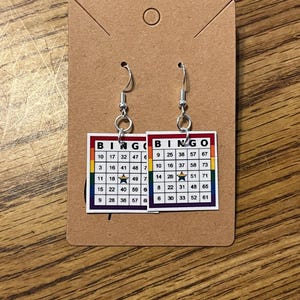 May include: A pair of Bingo card earrings. Each earring features a Bingo card design with a rainbow border and the word "BINGO" at the top. The cards have numbers in various colors and a silver fish hook.