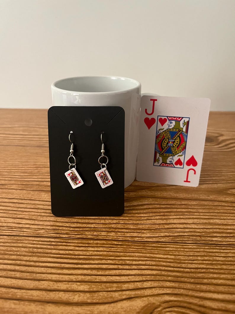 May include: A pair of dangle earrings with a black card design featuring the Queen of Hearts. The earrings are on a black card display.