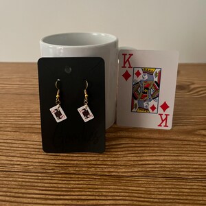 May include: A pair of gold earrings with a small playing card charm. The card is a black jack of diamonds. The earrings are displayed on a black card with the word "Earles" printed on it. A king of diamonds playing card is in the background.
