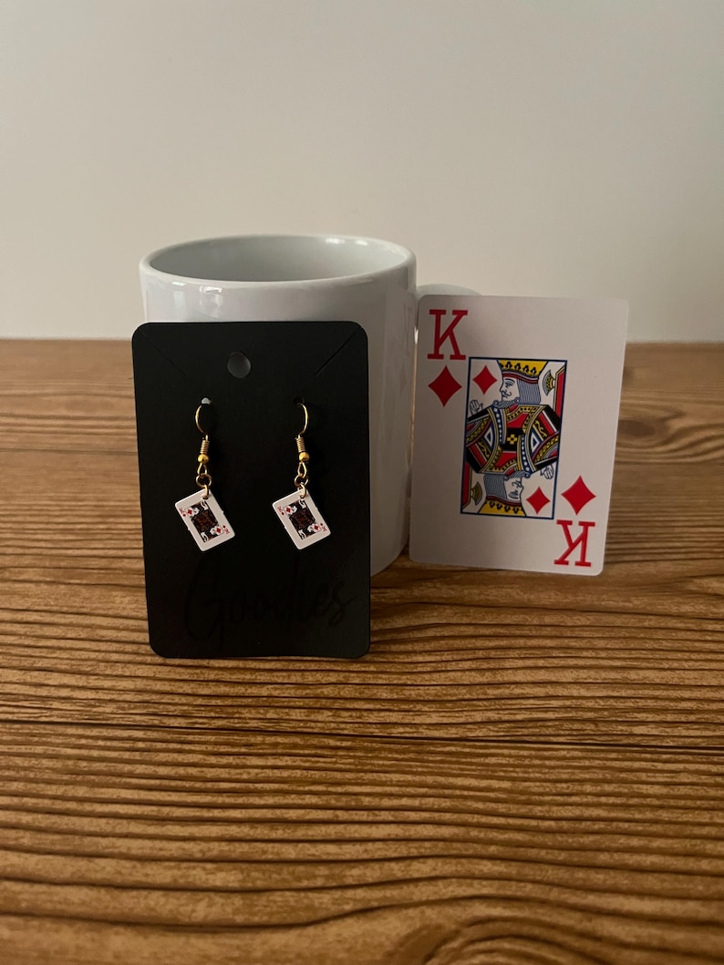 May include: A pair of gold earrings with a small playing card charm. The card is a black jack of diamonds. The earrings are displayed on a black card with the word "Earles" printed on it. A king of diamonds playing card is in the background.