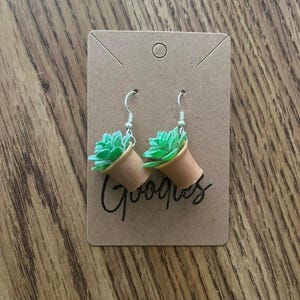 May include: A pair of dangle earrings featuring small potted plants with green leaves and brown pots. The earrings are on a brown card with the word "Gooques" written on it.
