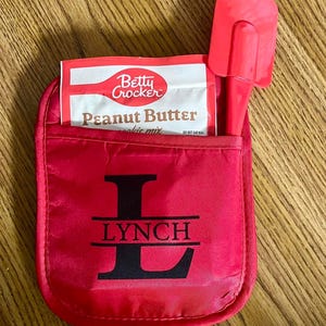 May include: Red oven mitt with the letter L and the name LYNCH printed on it. A package of Betty Crocker Peanut Butter Cookie Mix and a red spatula are inside the mitt.
