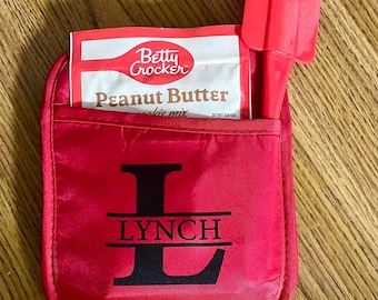 May include: A red fabric oven mitt with a pocket containing a Betty Crocker Peanut Butter cookie mix packet and a red silicone spatula. The front of the mitt features a large black letter L with the name LYNCH printed across the center.
