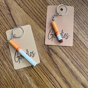 May include: Two brown card stock tags with the word "Goodies" written in black cursive. Each tag has a silver key ring attached to a small, realistic-looking cigarette keychain.