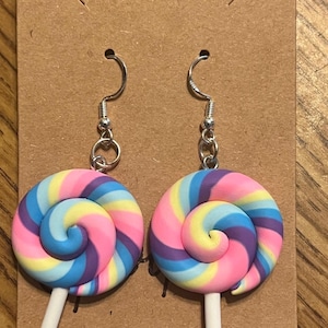 May include: A pair of silver dangle earrings with a pink, yellow, blue, and purple swirl lollipop charm. The card behind the earrings says "Unicorn".