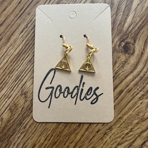 May include: Gold-colored earrings featuring a triangle design with a circle in the center, displayed on a tan card. The card has the word "Goodies" in a handwritten style. The earrings are on a wooden surface.