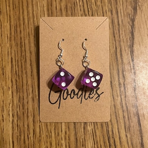 May include: A pair of dice earrings with silver hooks. The dice are transparent purple with white dots. The earrings are displayed on a brown card with the word "Goodies" in black script.