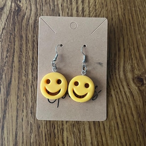 May include: A pair of yellow smiley face earrings featuring circular charms with cut-out eyes and a wide grin. The charms are attached to silver-toned metal fishhook ear wires and are displayed on a rectangular brown cardboard jewelry card.