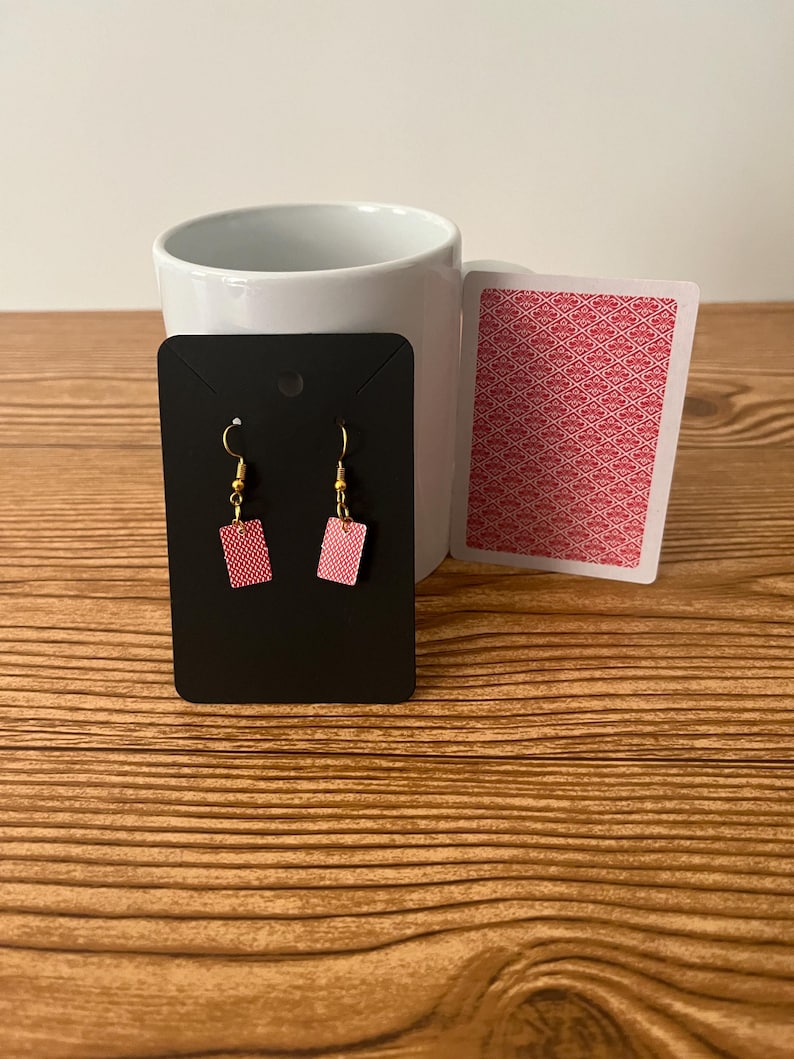 May include: A pair of red playing card earrings on a black display card. The earrings are shaped like a playing card and have a gold hook.