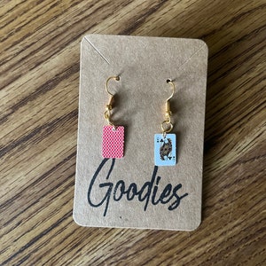 May include: A pair of gold earrings with a red playing card and a Queen of Spades playing card charm. The earrings are displayed on a brown card with the word "Goodies" written on it.