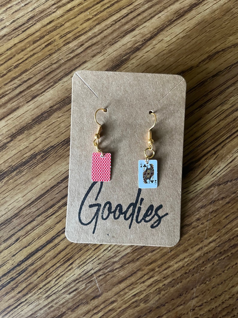 May include: A pair of gold earrings with a red playing card and a Queen of Spades playing card charm. The earrings are displayed on a brown card with the word "Goodies" written on it.