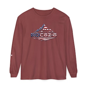 May include: A long-sleeve crew neck t-shirt in a muted brick red color. The chest features a graphic of a mountain range filled with the American flag pattern, positioned above the text ROC62:6. A small white star is printed on the left sleeve.