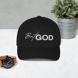 Op de afbeelding: Zwarte baseballpet met een wit geborduurd ontwerp dat "But GOD" zegt.