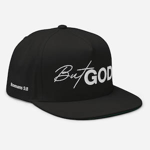 Op de afbeelding: Zwarte baseballpet met platte klep met de woorden "But God" in witte letters over de voorkant. De tekst "Romeinen 5:8" staat op de zijkant. De pet heeft een groene onderkant.