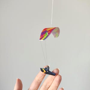 May include: A miniature paraglider mobile with a colorful rainbow-patterned canopy and a dark blue harness with yellow accents. The mobile is suspended by a thin white string, creating a floating effect.