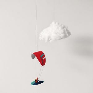 May include: A hanging mobile featuring a small paraglider with a red canopy and a dark blue board, suspended by thin white strings alongside a single, fluffy white cloud made of soft material, creating a suspended aerial scene.
