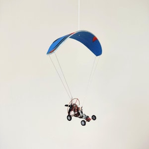 May include: A miniature paraglider model with a blue and red canopy. The model features a small cart with wheels and a propeller. The paraglider is suspended by thin white strings against a plain background.
