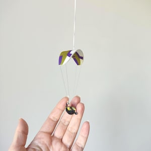 May include: A miniature paraglider with a purple, green, and white canopy is suspended in the air. The paraglider is attached to a small figure. The image is set against a plain background.