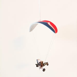 May include: A small, red, white and blue hang glider with a black engine and propeller hanging from a white string against a white background.