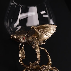 Op de afbeelding: Een glazen kelk met een gouden schorpioen- en vleermuisontwerp. De staart van de schorpioen wikkelt zich om de kelk, en de vleugels van de vleermuis zijn uitgespreid. De vleermuis heeft rode ogen.