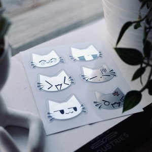 May include: A sheet of six white cat stickers with different facial expressions. The stickers are on a white background and have a glossy finish.