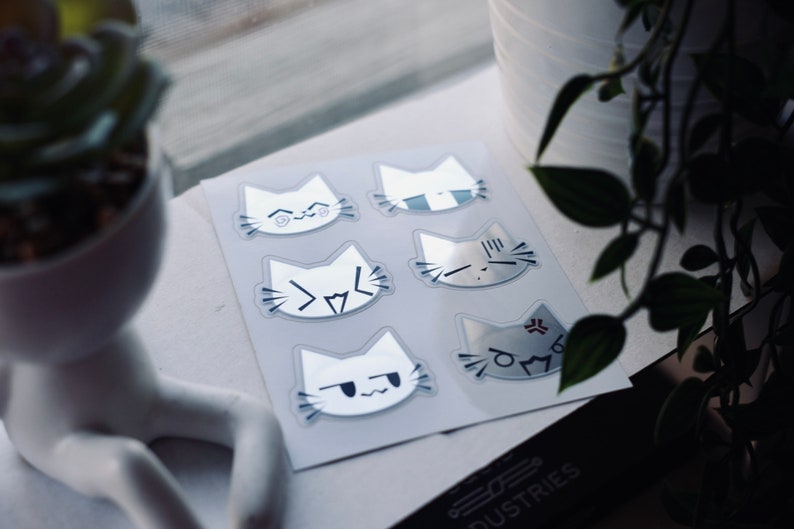 May include: A sheet of six white cat stickers with different facial expressions. The stickers are on a white background and have a glossy finish.