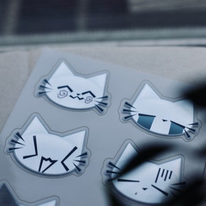 May include: A sheet of stickers featuring six white cat faces with different expressions. The cat faces are outlined in black and have various black markings on their faces.