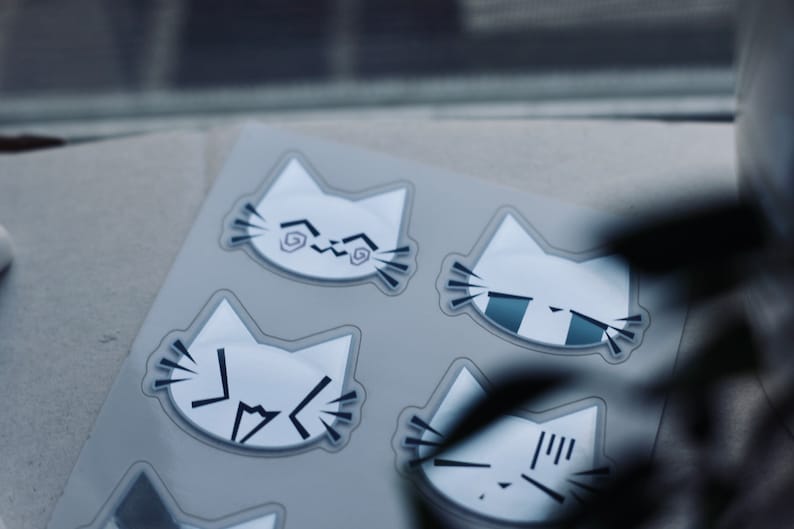 May include: A sheet of stickers featuring six white cat faces with different expressions. The cat faces are outlined in black and have various black markings on their faces.
