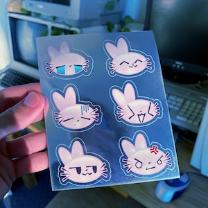 Könnte beinhalten: Ein Bogen Aufkleber mit sechs rosa Hasenillustrationen mit verschiedenen Gesichtsausdrücken. Die Hasen haben lange Ohren, Schnurrhaare und sind schwarz umrandet. Die Ausdrücke reichen von fröhlich bis wütend, auf einem reflektierenden blauen Hintergrund.