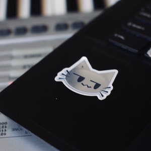 May include: A gray cat sticker with a grumpy expression, placed on a black laptop. The laptop is sitting on top of a gray keyboard.
