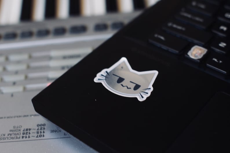 May include: A gray cat sticker with a grumpy expression, placed on a black laptop. The laptop is sitting on top of a gray keyboard.