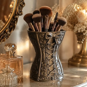 May include: A dark metallic grey makeup brush holder shaped like a corset with intricate scrollwork, a central lace-up design, and a bow detail at the top. The holder is filled with various makeup brushes featuring brown bristles and metallic handles.
