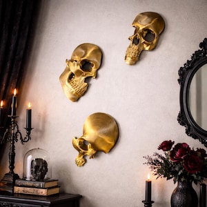 May include: Three gold-colored skull wall decorations are mounted on a light gray wall. The skulls are in different positions, creating a dynamic display. The scene includes a black candle holder, a mirror, and a vase of red flowers, suggesting a gothic or Halloween theme.