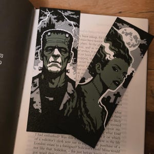 Op de afbeelding: Twee zwart-witte bladwijzers met illustraties van Frankenstein en de bruid van Frankenstein. De bladwijzer van het monster heeft een grijze achtergrond met bliksem. De bladwijzer van de bruid heeft een volle maan en een kerkhofachtergrond.