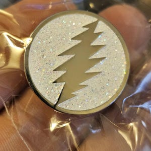 May include: A white glitter pin with a gold lightning bolt design. The pin is circular and has a shiny finish.