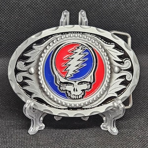 May include: Oval-shaped silver belt buckle featuring a central design of a skull with a lightning bolt, surrounded by red and blue circles. The buckle has a decorative border with floral and scrollwork details.