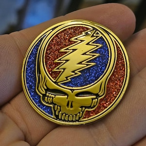 May include: Gold-colored metal pin with a red and blue glitter background. The pin features a stylized skull with a lightning bolt design inside.